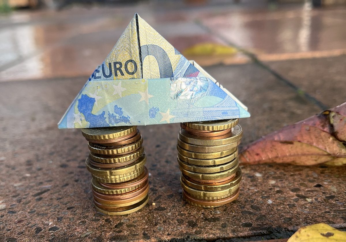 Das Symbolbild zum Thema Wohngeld zeigt zwei Stapel mit Euro-Münzen, auf denen ein zu einem spitzen Dach gefalteter 20-Euro-Schein liegt. Das Ganze sieht aus wie ein Haus.